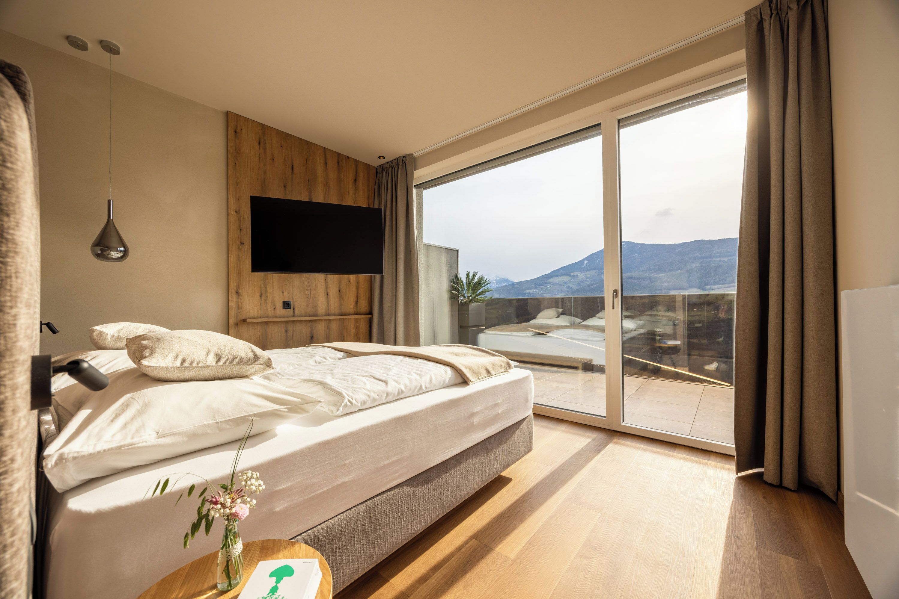 A modern bedroom with a large bed, a flat-screen TV, and a balcony with a view of the mountains.