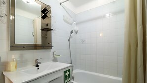 Ein Badezimmer mit einem weißen Waschbecken, einem Spiegel und einer Dusche. An der Wand befindet sich ein grünes Notzeichen.