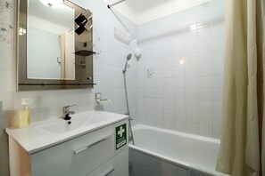 Un baño con lavamanos blanco, espejo y ducha. Hay un letrero de emergencia verde en la pared.
