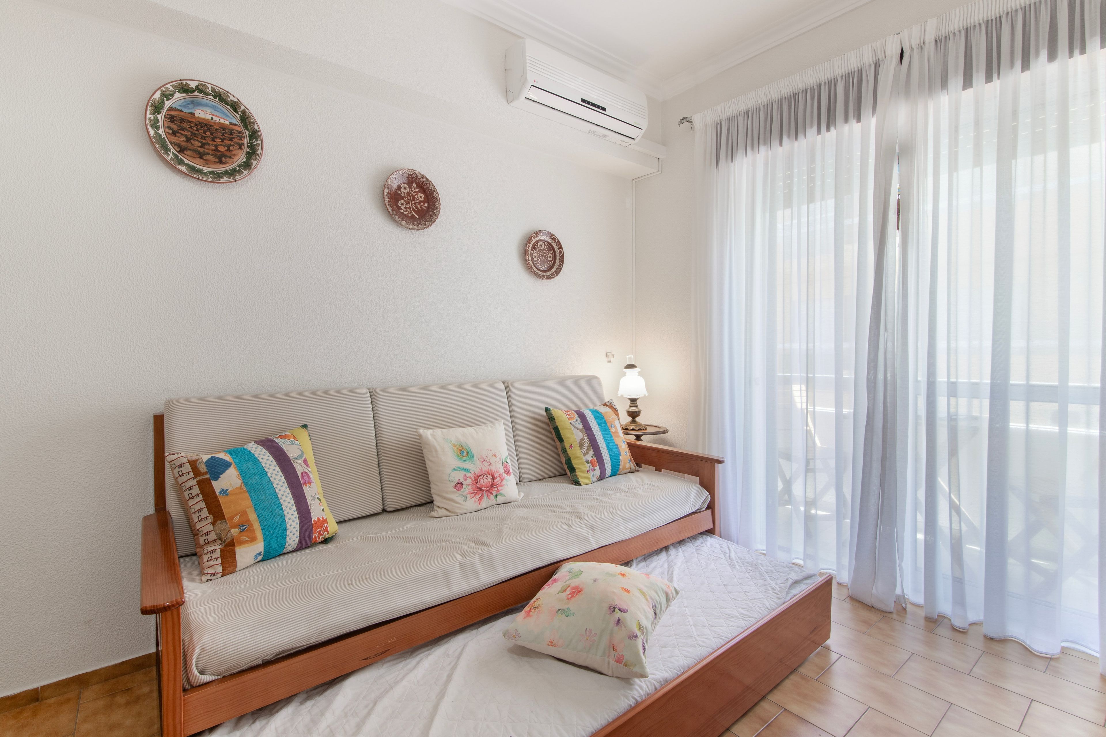 A room with a sofa bed, a bedside table with a lamp, and a window with sheer curtains.