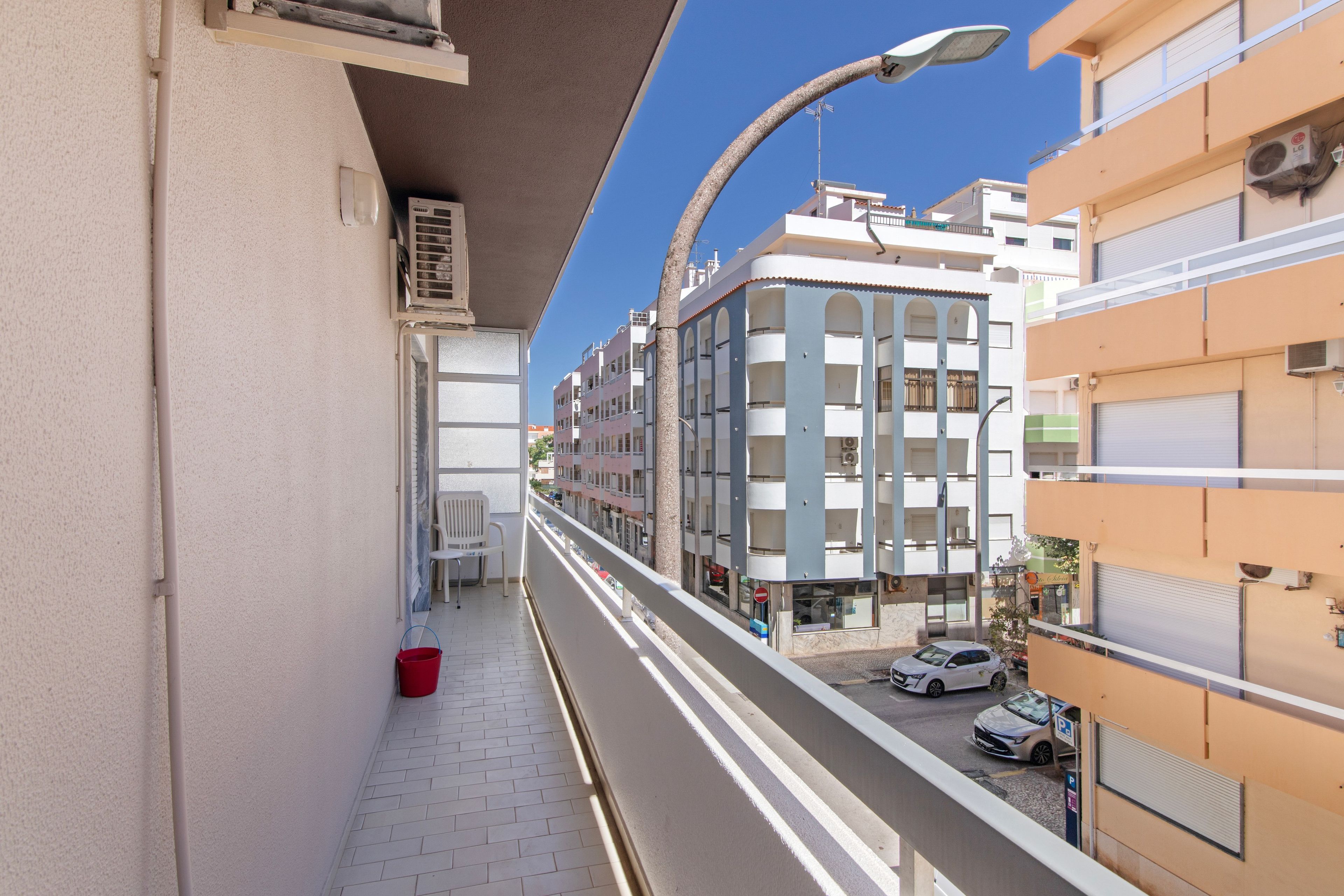 A narrow urban street with buildings on either side, a clear sky, and parked cars.