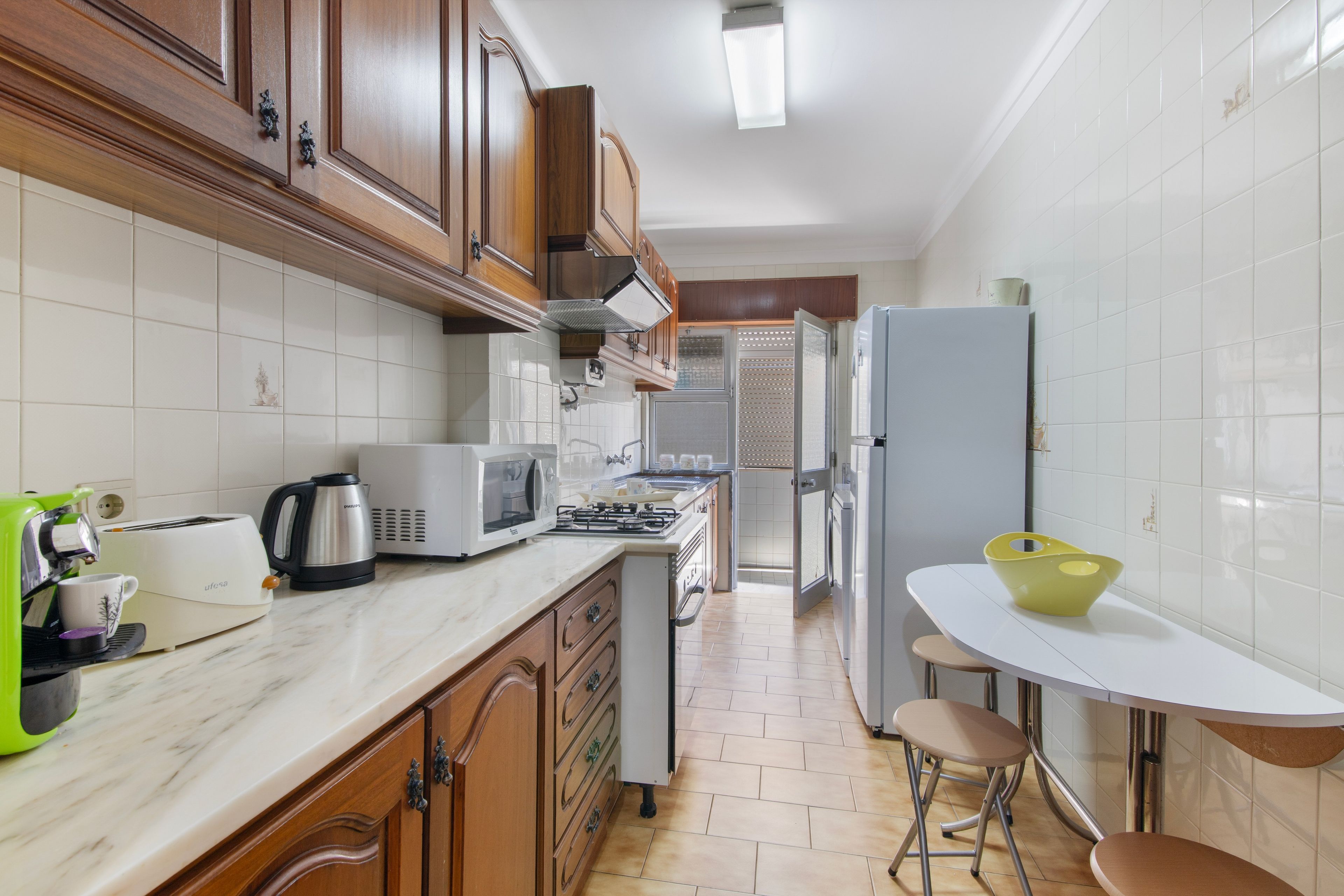 A kitchen with wooden cabinets, a white countertop, a microwave, a kettle, and a small dining table with stools.
