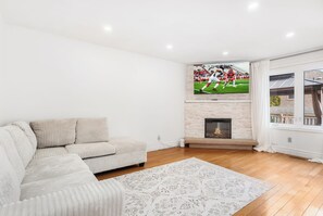 A modern living room with a sectional sofa, a fireplace, and a large TV screen displaying a football game. - Executive 5BR Rose Bloom Haven with Private Deck (Mississauga)