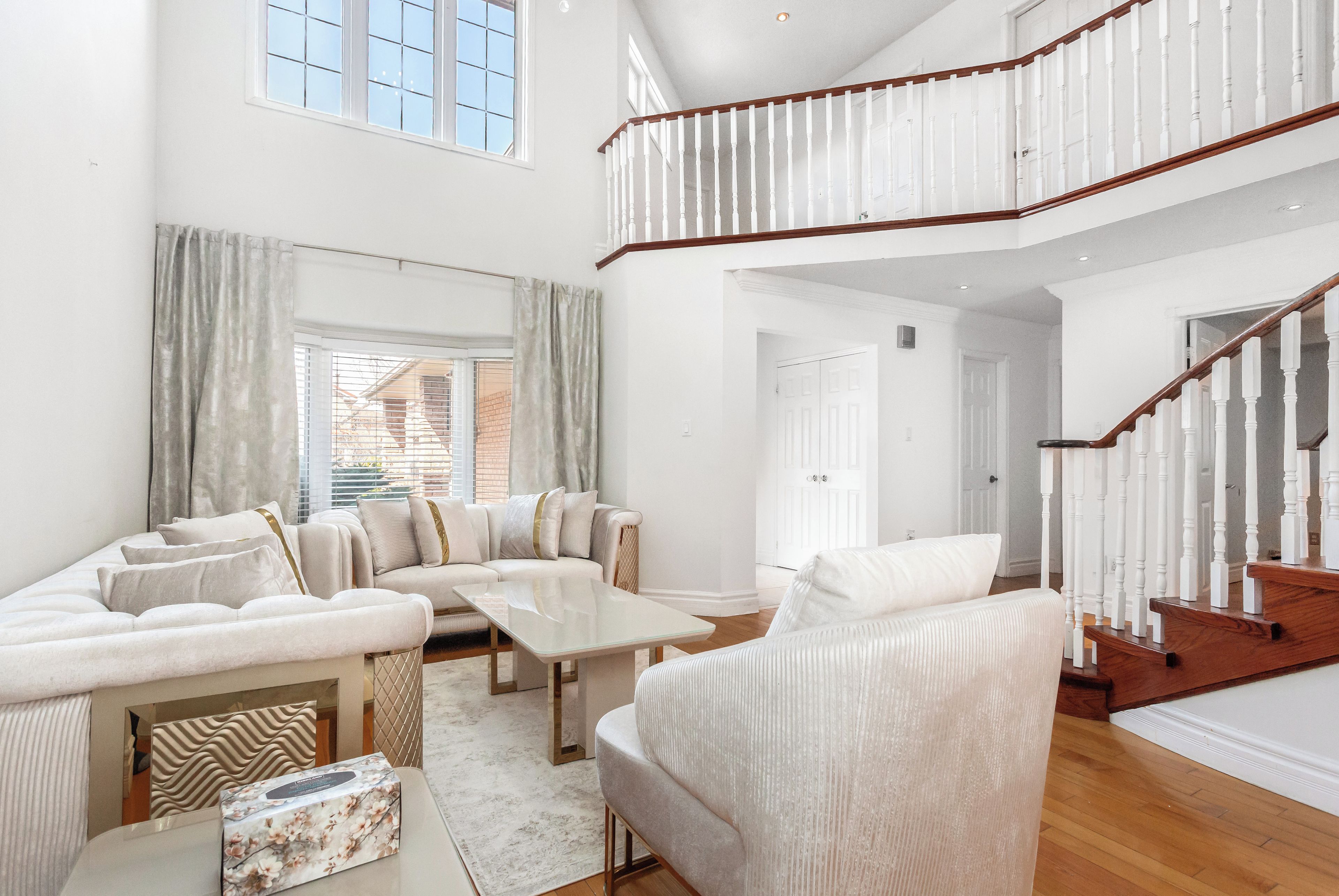 A spacious living room with a high ceiling, a staircase with white balusters, and a view through the window.