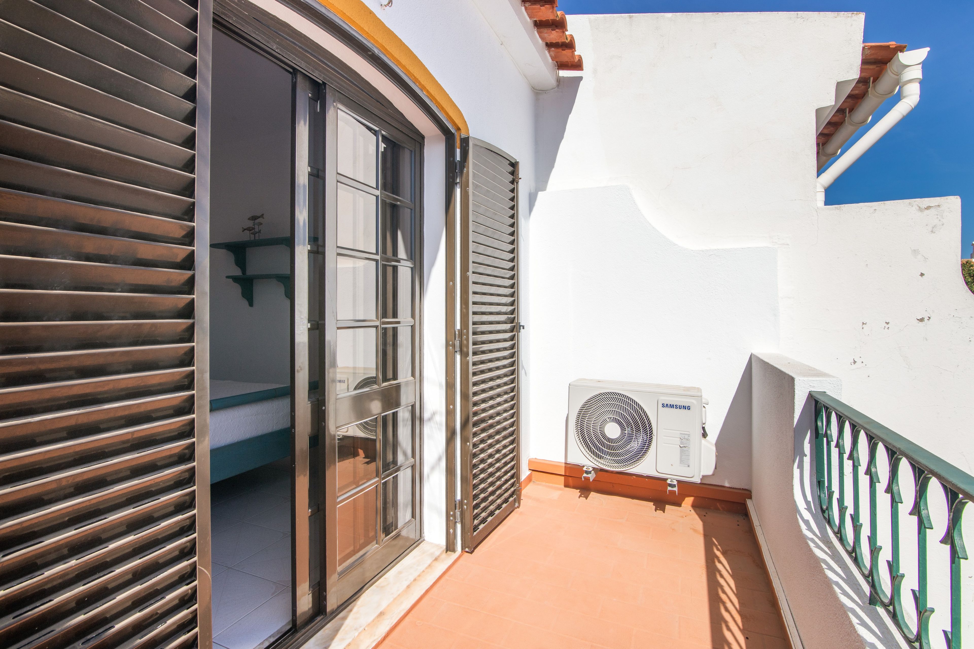 Un estrecho pasillo exterior con puerta corredera, aire acondicionado en la pared y suelo de baldosas.