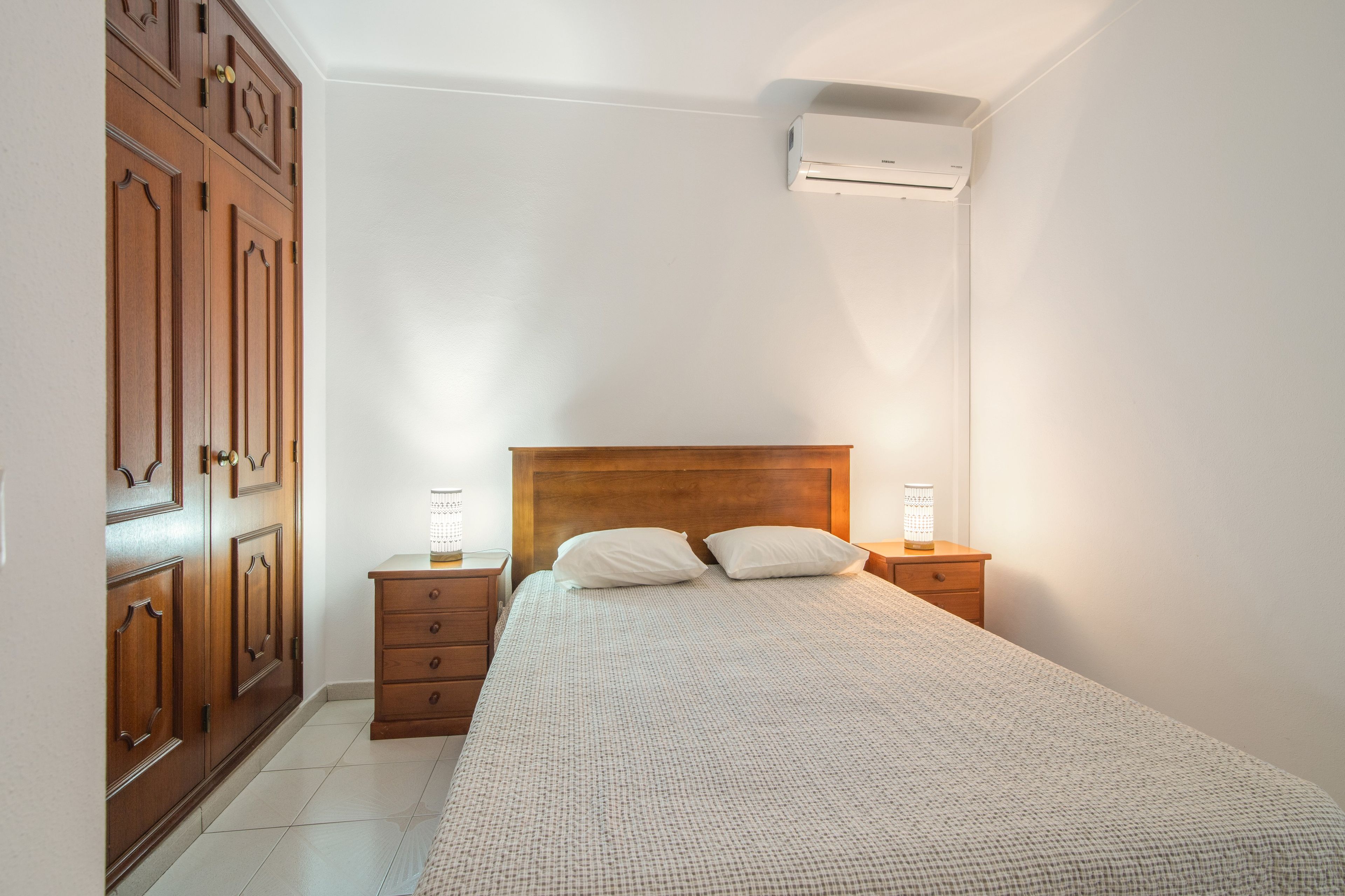 A bedroom with a wooden bed, two bedside tables with lamps, a wardrobe, and an air conditioning unit.