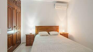 A bedroom with a wooden bed, two bedside tables with lamps, a wardrobe, and an air conditioning unit.