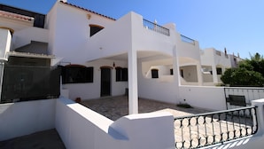 Un edificio blanco de dos pisos con techo de tejas, un balcón con barandillas negras y un patio empedrado.