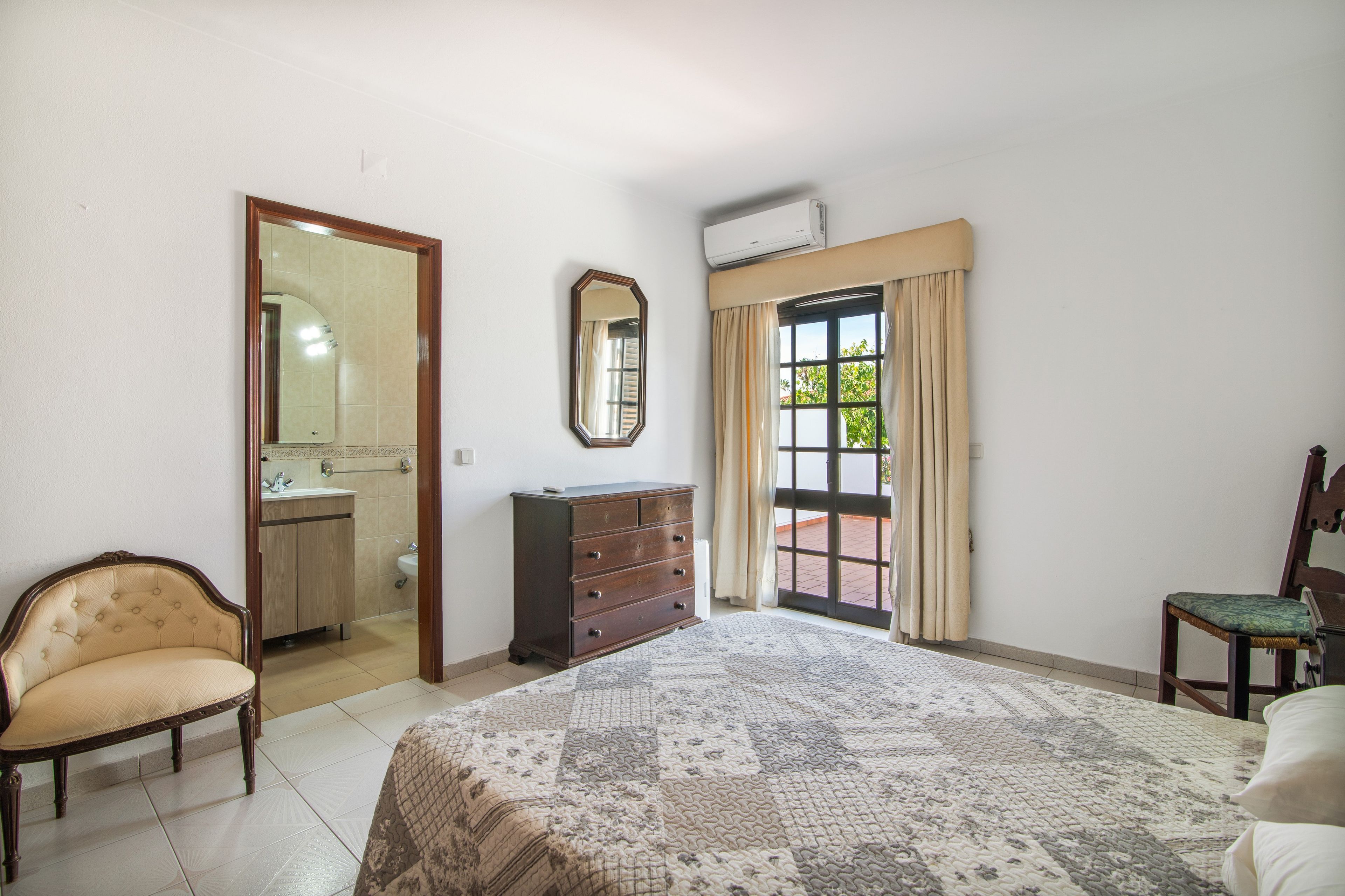 Un dormitorio con cama, cómoda, silla, espejo y baño visible a través de una puerta abierta.