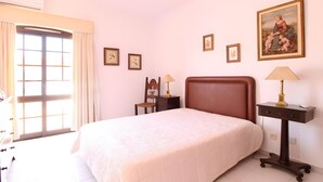 Un dormitorio con cama, mesitas de noche, una silla y una ventana con cortinas.