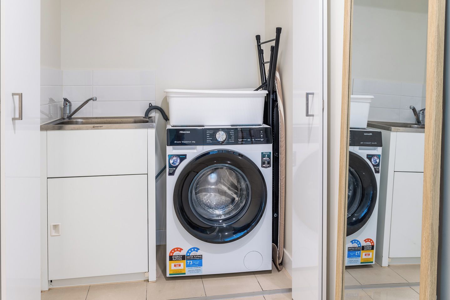 A compact, modern laundry room with a front-loading washing machine, a sink, and a mirror.