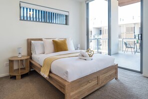 A bedroom with a wooden bed, white bedding, a bedside table with a lamp, and a view of an outdoor balcony.