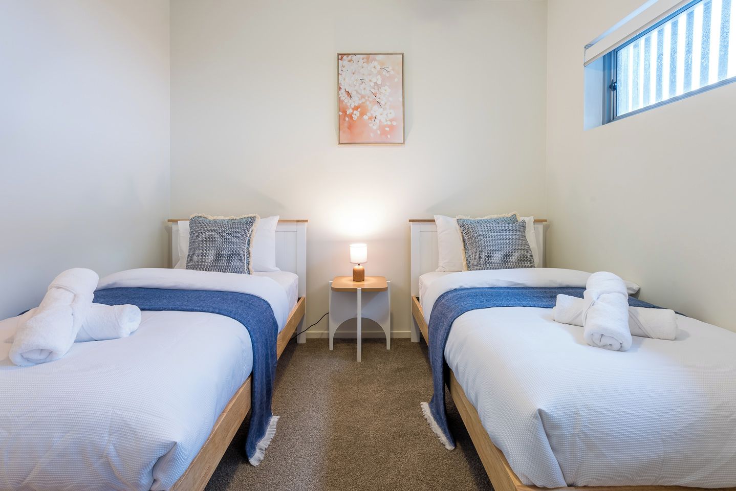 A hotel room with two single beds, a small bedside table with a lamp, and a window with blinds.