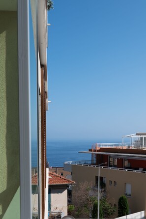 A coastal view from a balcony with a clear sky, a building with a red roof, and a distant view of the sea. - Cozy 1-bedroom Apartment With A C Garage Near (Beausoleil)