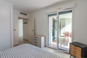 Une chambre à coucher comprenant un lit, une commode, une porte-fenêtre coulissante donnant sur un balcon, et une petite table avec des chaises.