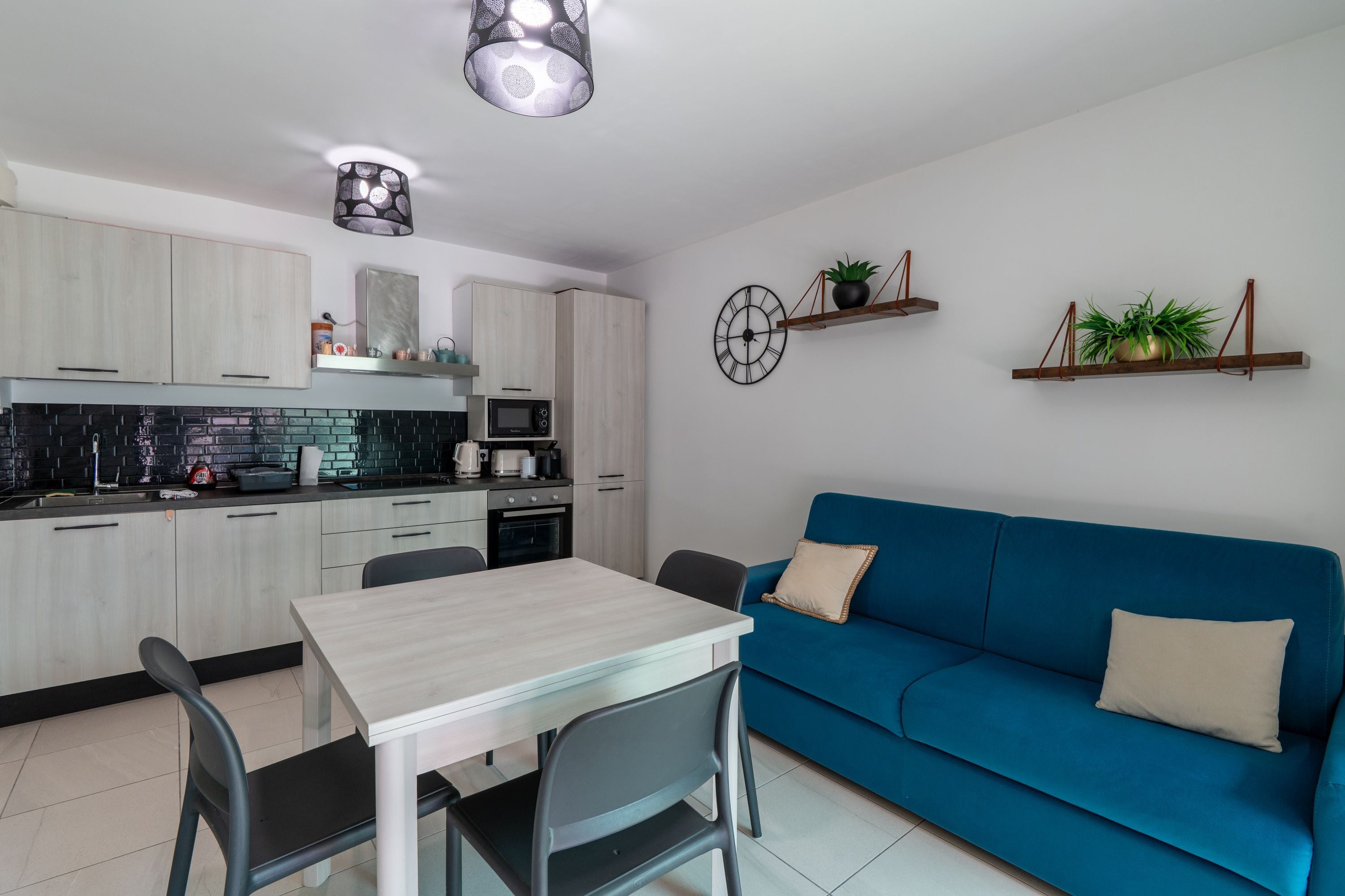 Eine moderne Küche mit Esstisch und Stühlen, einem blauen Sofa und zwei an der Wand befestigten Regalen mit Pflanzen.