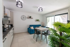 A modern kitchen with a dining area, a blue sofa, and a table with chairs. There are ceiling lights, a clock, and shelves with plants. - Cozy 1-bedroom Apartment With A C Garage Near (Beausoleil)
