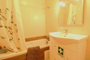 Un baño con bañera, lavamanos y espejo. Hay un perchero y un cartel de primeros auxilios en la pared.