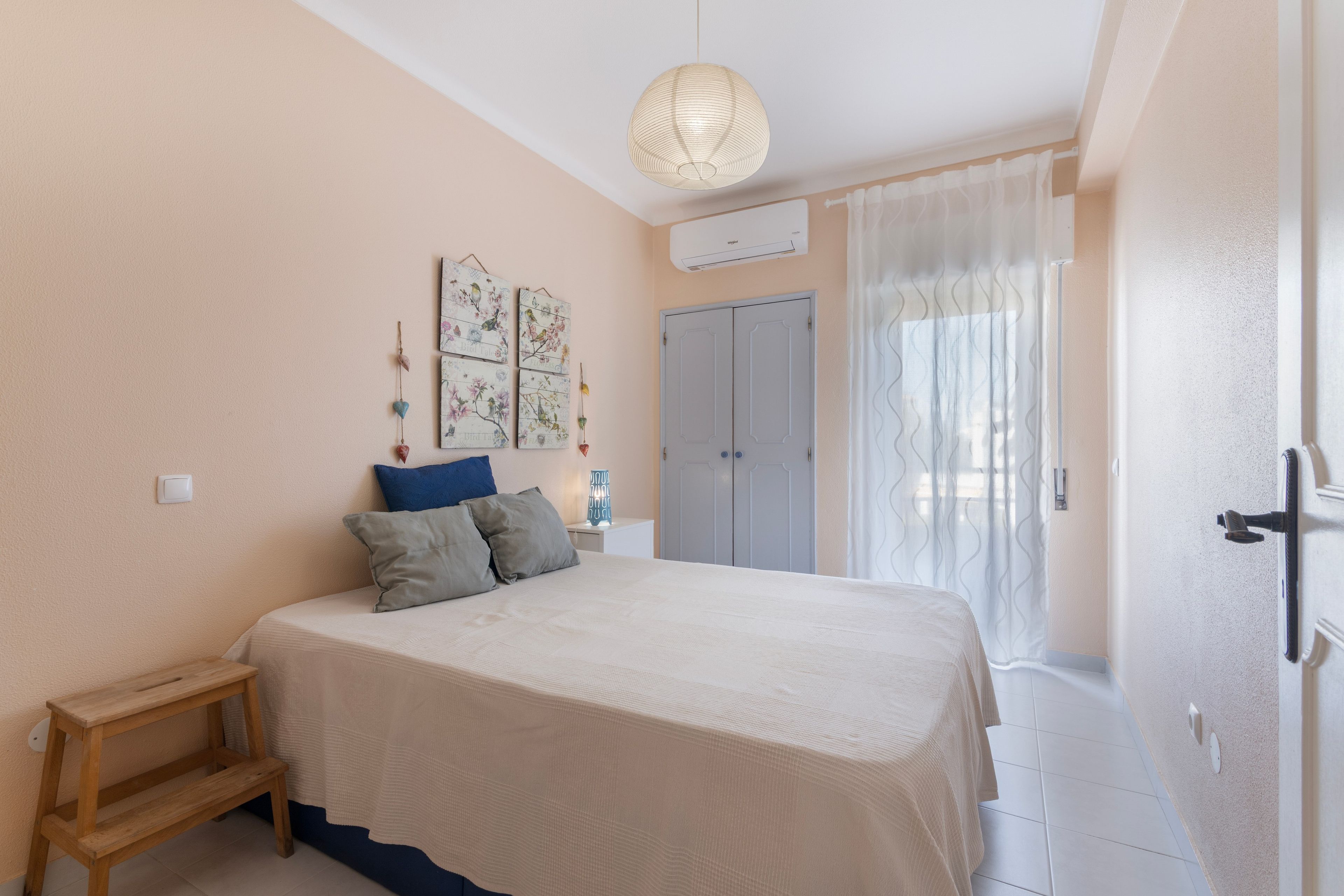 Une chambre à coucher comprenant un lit, une table de chevet, un luminaire suspendu, une armoire et une fenêtre avec des rideaux.