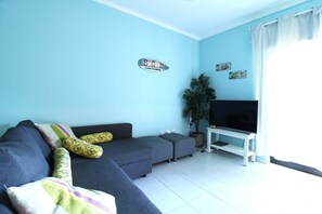 Un salón con un sofá gris, un televisor de pantalla plana sobre un mueble, una planta en maceta y una pared con un cartel de temática playera.