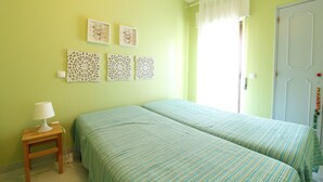 A room with two beds, a bedside table with a lamp, decorative wall art, and a door with a curtain. - Holiday Home 'Mantamar Beach Ap' with Balcony, Wi-Fi and Air Conditioning (Vila Nova de Cacela)