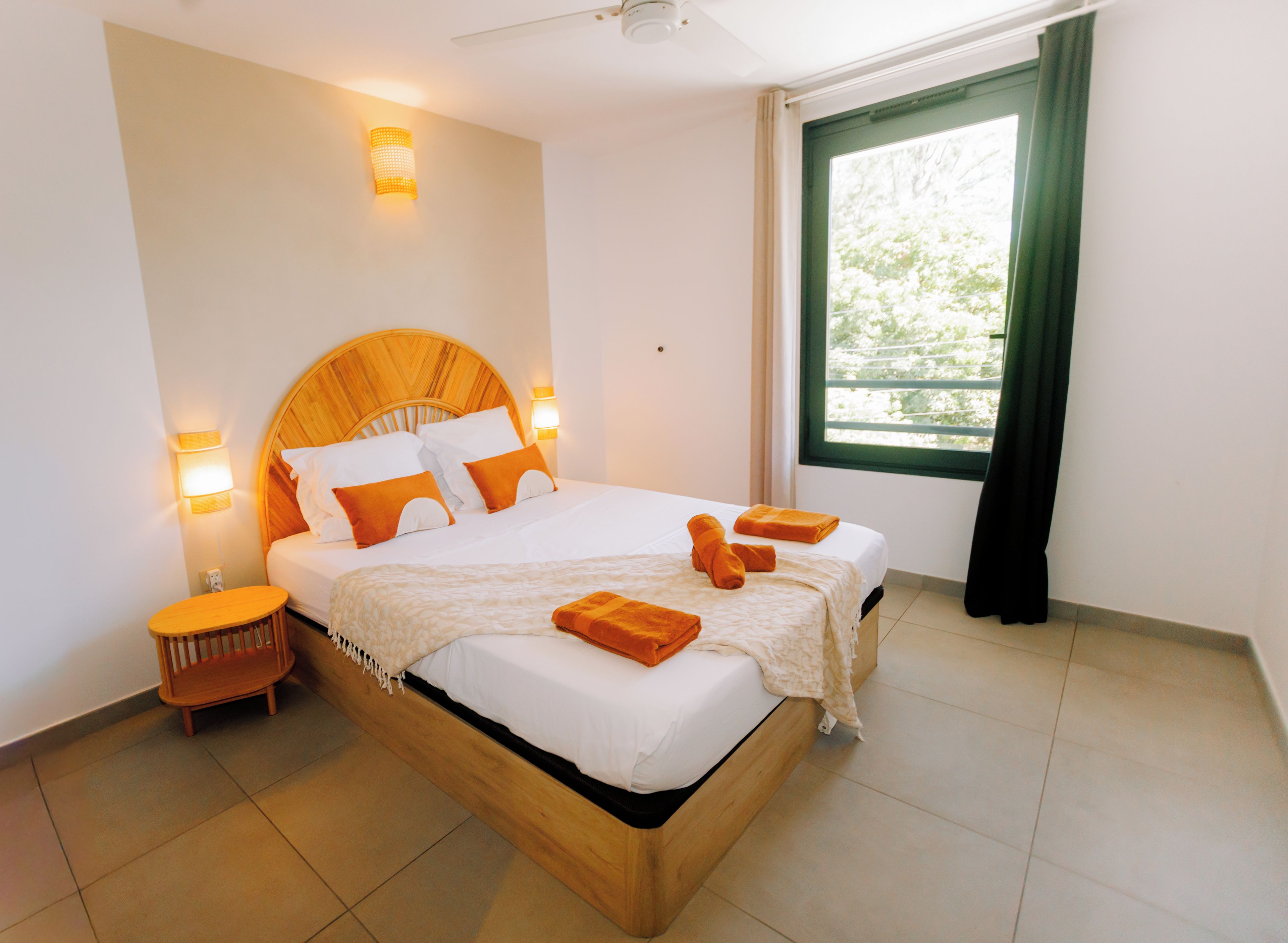 A hotel room with a wooden headboard, a bed with white bedding and orange pillows, a nightstand, and a window with curtains.