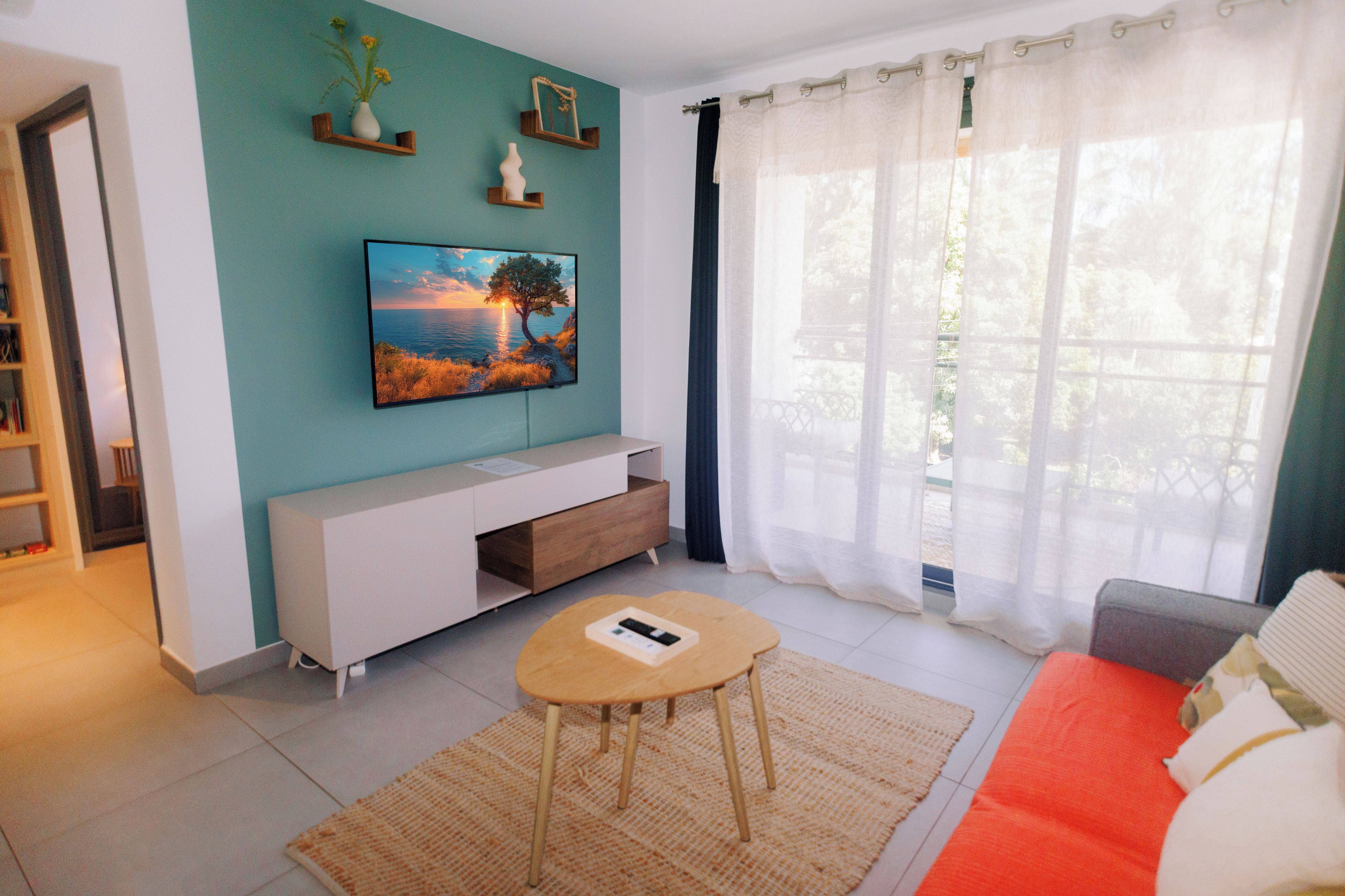 Een woonkamer met een wandtelevisie, een bank, een salontafel en een raam met dunne gordijnen.