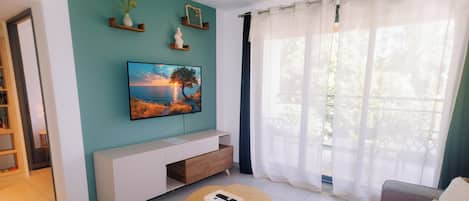 Una sala de estar con televisor de pantalla plana montado en la pared, un sofá, una mesa de centro y una ventana con cortinas translúcidas.