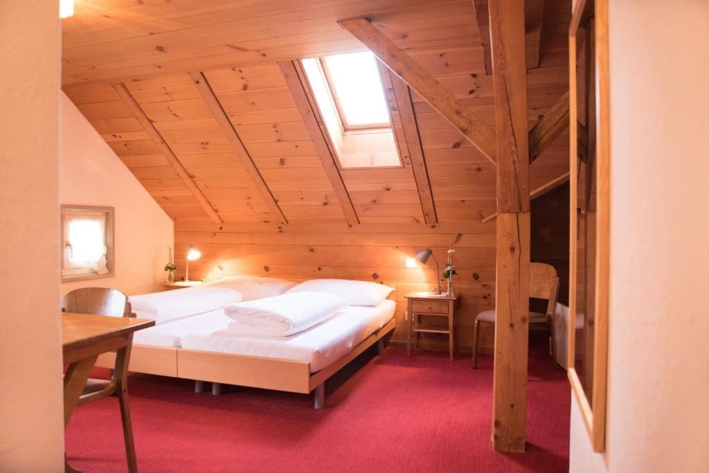 A bedroom with a sloped ceiling, a skylight, a bed with white linens, a bedside table with a lamp, and a chair.