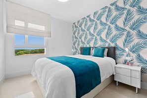 A bedroom with a bed, a nightstand, a lamp, and a window with a view of greenery. - EMERALD GREEN 2477 SAN ROQUE (San Roque)