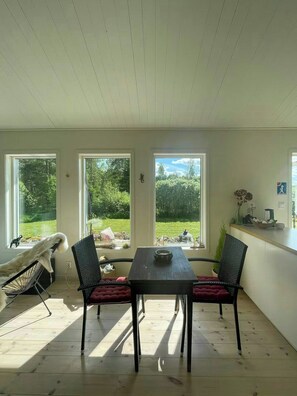 Un espace repas comprenant une table et des chaises, de grandes fenêtres donnant sur un paysage verdoyant et un plafond avec des poutres en bois.