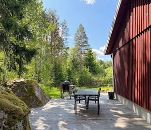 Une terrasse en bois avec une table et des chaises, entourée d’une zone boisée et d’un bâtiment rouge.