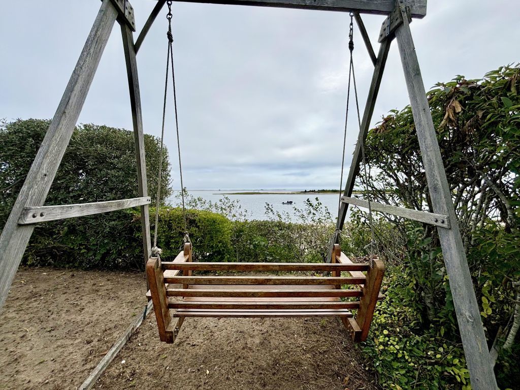 Um conjunto de baloiço de madeira com um banco, rodeado por vegetação e com vista para um corpo de água.