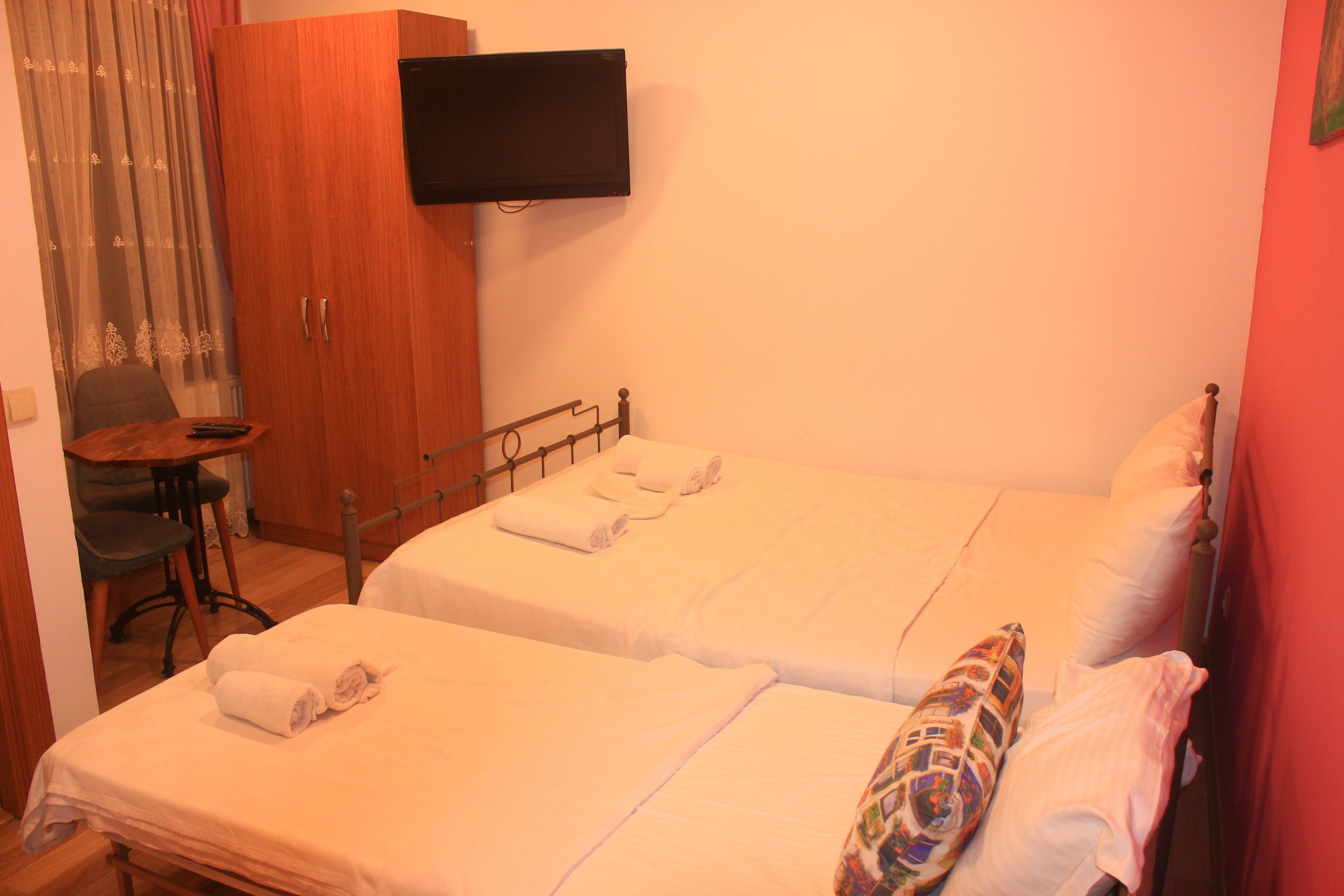 Habitación de hotel con dos camas, un televisor montado en la pared, un armario de madera y una mesita con una silla.