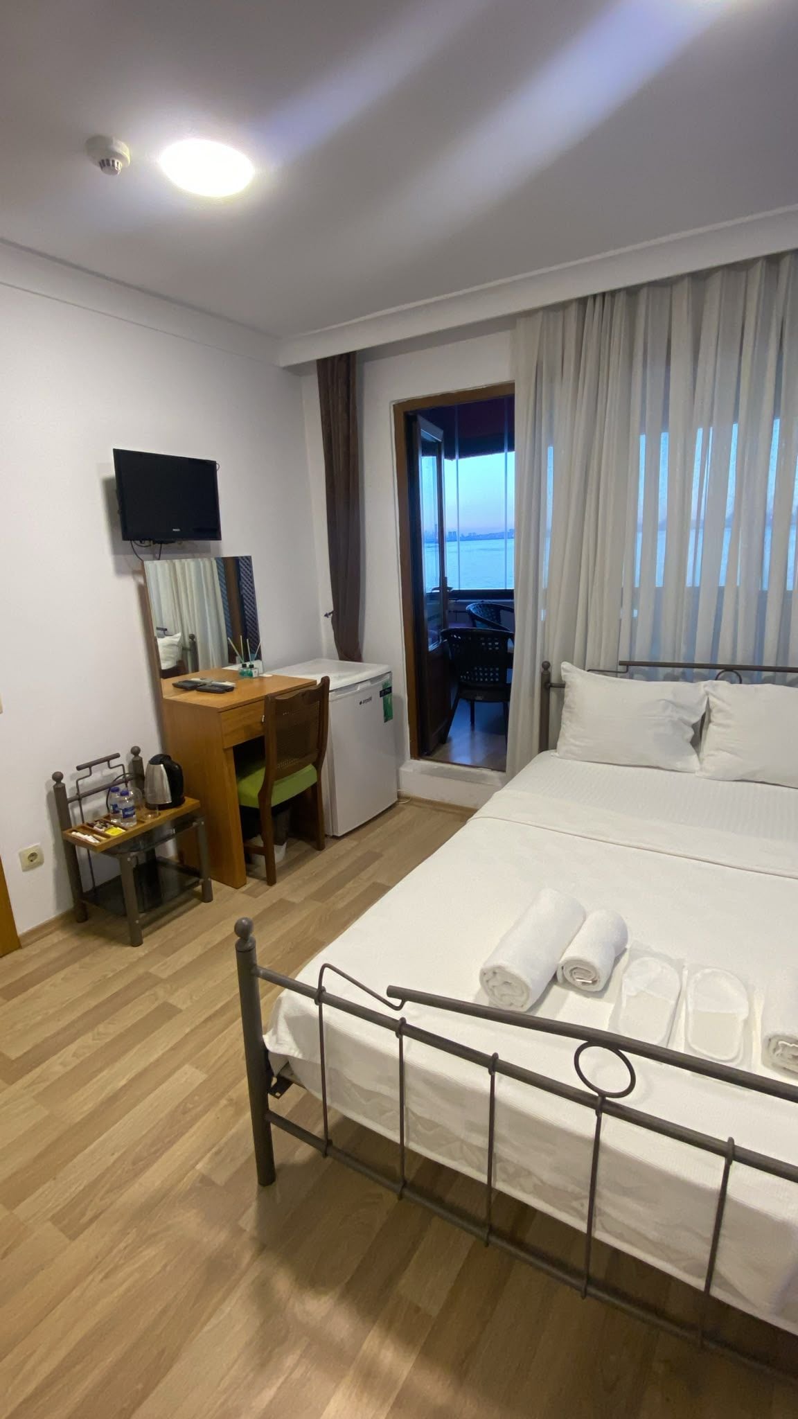A hotel room with a bed, a desk, a chair, a small refrigerator, a television, and a balcony with a view.