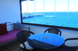 Un balcón con vista al mar y un barco, una mesa redonda con un vaso y sillas de mimbre.