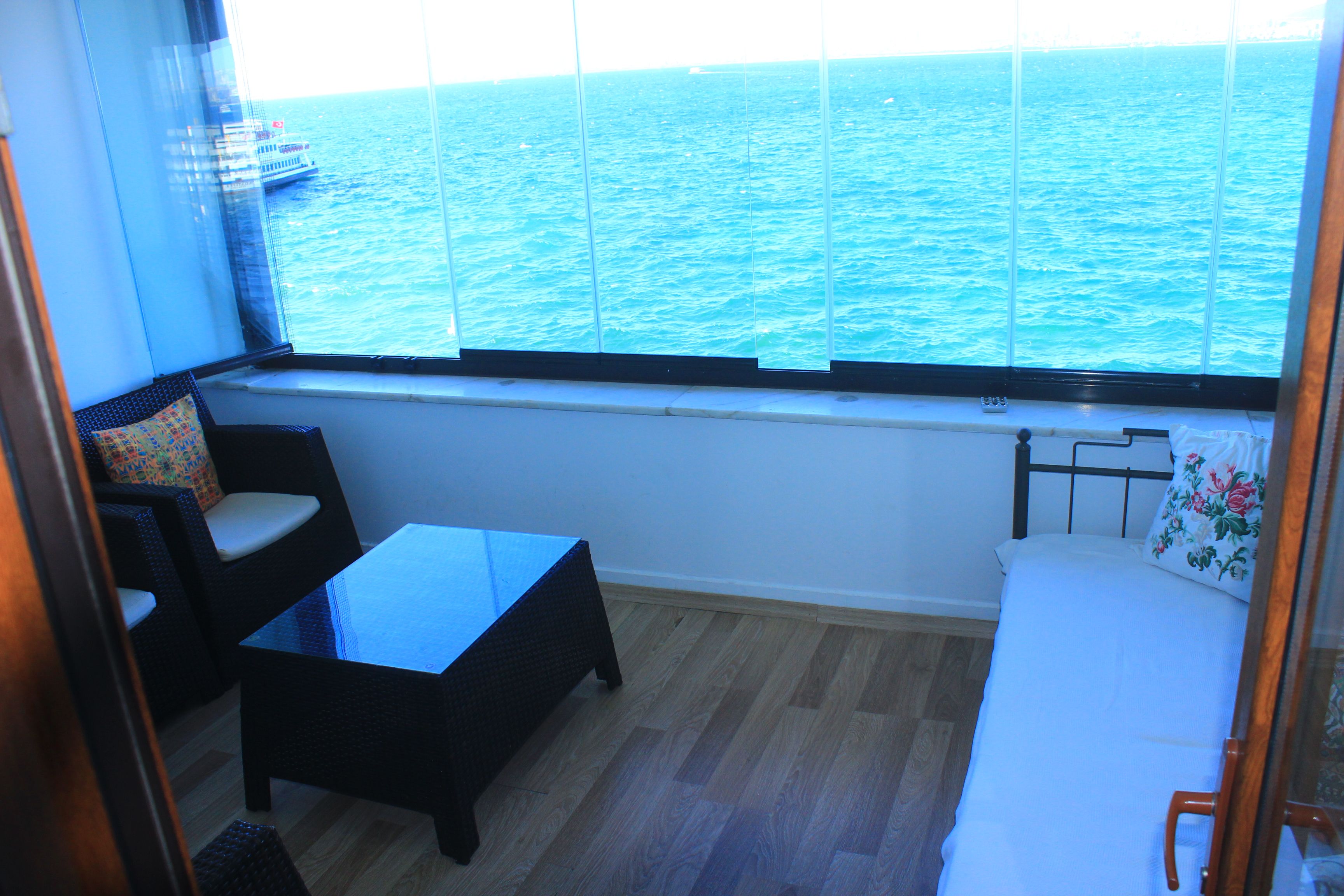 Un pequeño balcón con vista al mar, una silla de mimbre, una mesita y una cama con un cojín floral.