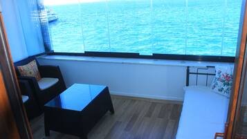 Un pequeño balcón con vista al mar, una silla de mimbre, una mesita y una cama con un cojín floral.