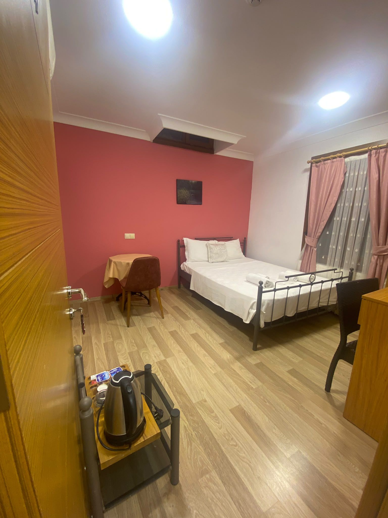 Habitación de hotel con cama, una mesita, una silla, un hervidor de agua y un ventanal con cortinas.