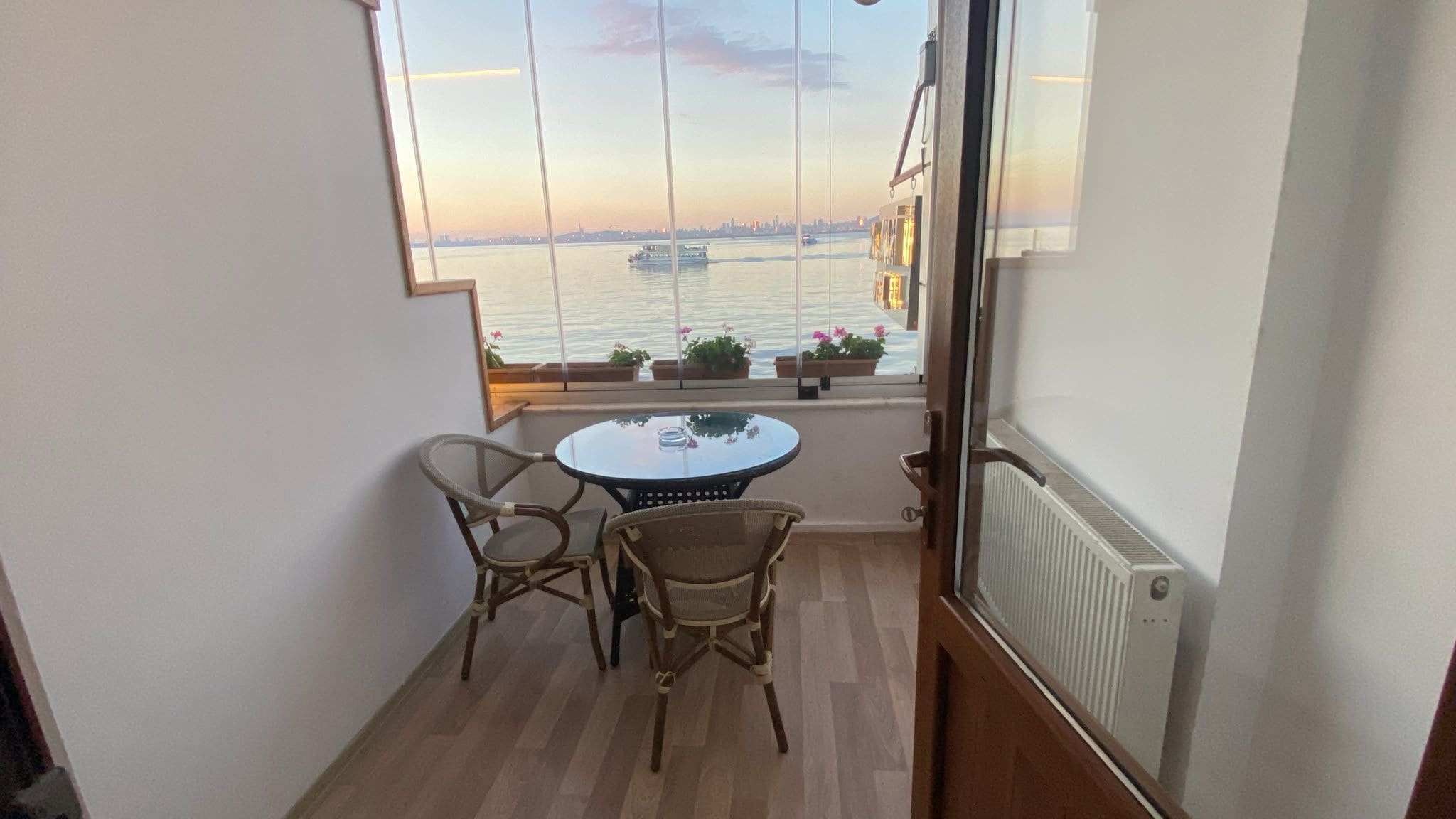 Un balcón con mesa y sillas, con vista a una masa de agua y un horizonte urbano distante.