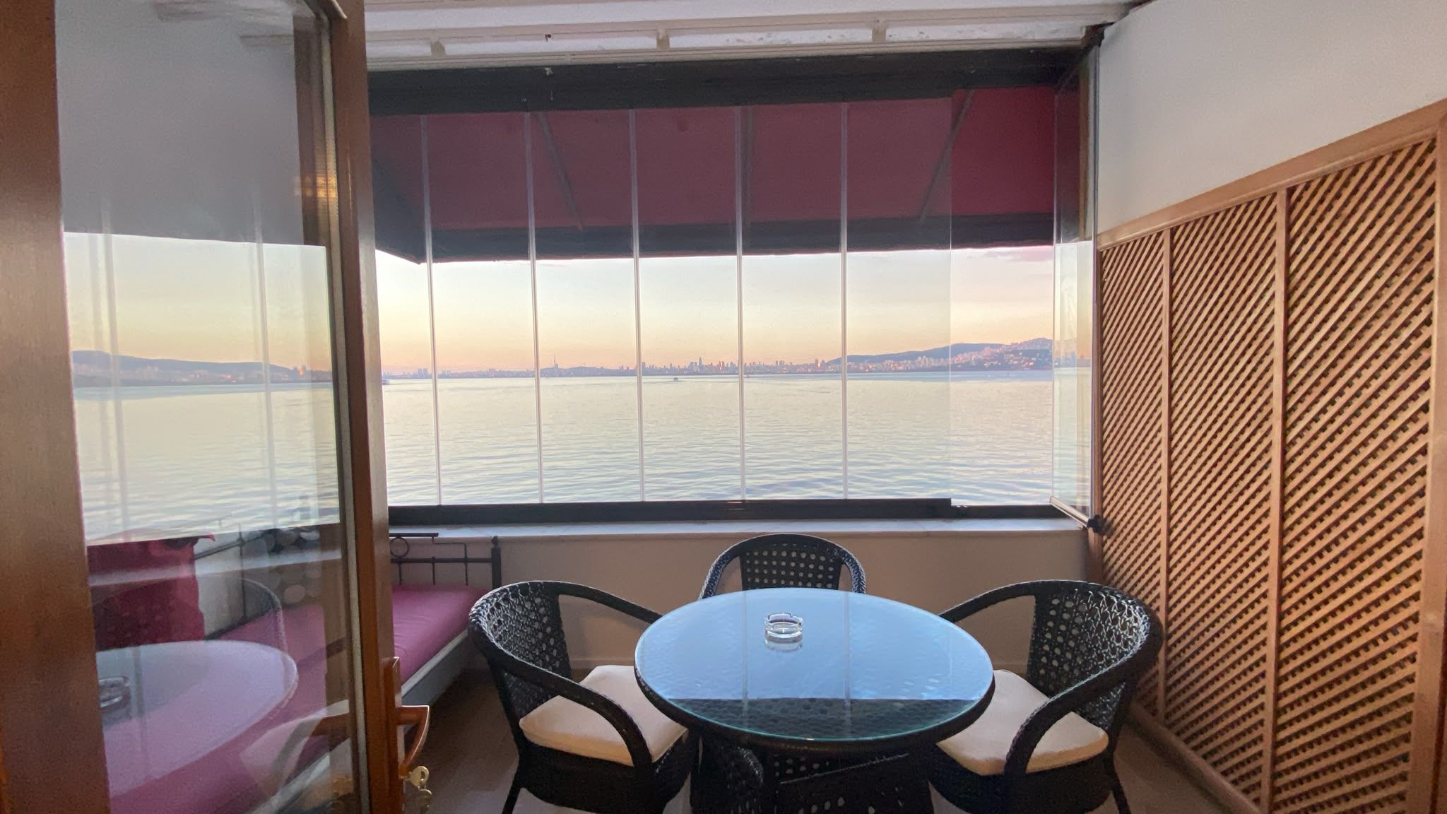 Un balcón con puerta de vidrio, una mesa redonda con sillas y vista a una masa de agua con tierra a lo lejos.
