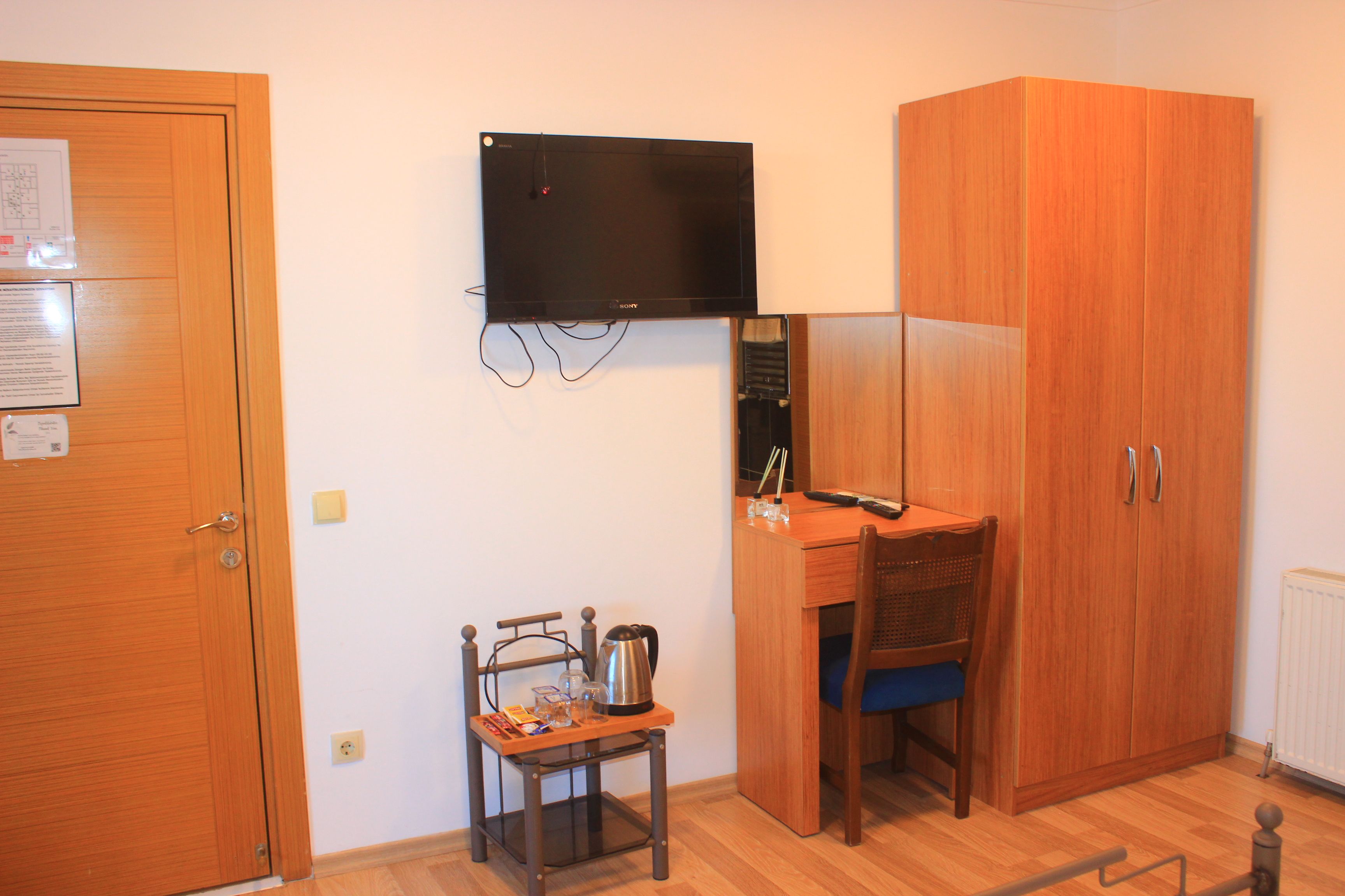 Ein Zimmer mit einem Holzkoffer, einem Schreibtisch mit Stuhl, einem kleinen Tisch mit Wasserkocher und einem an der Wand montierten Fernseher.