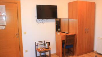 Ein Zimmer mit einem Holzkoffer, einem Schreibtisch mit Stuhl, einem kleinen Tisch mit Wasserkocher und einem an der Wand montierten Fernseher.