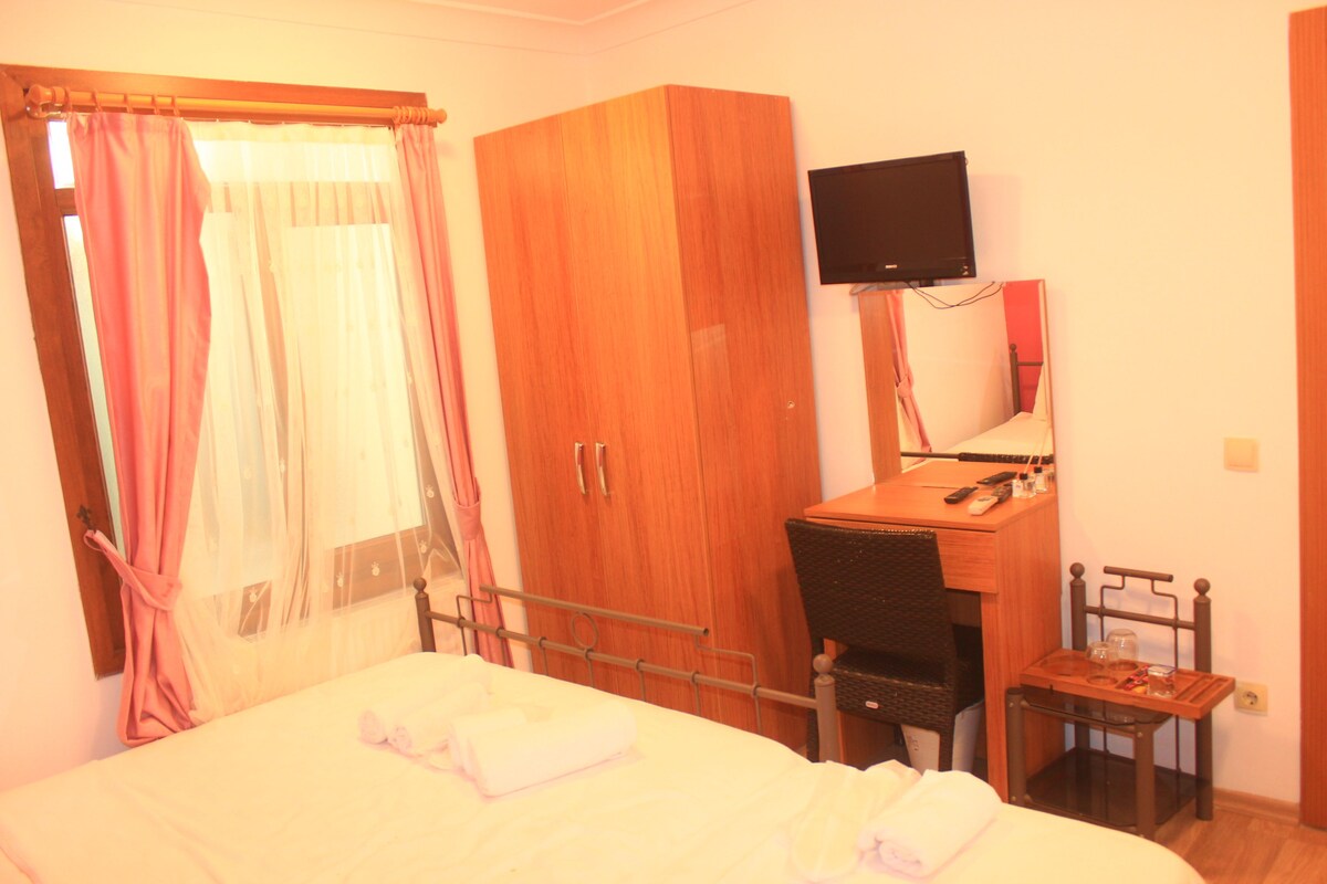 A bedroom with a bed, a wooden wardrobe, a desk with a chair, a television mounted on the wall, and a small table with items on it.