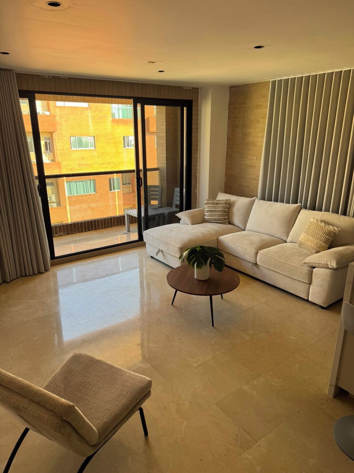Una sala moderna con un sofá, una mesa de centro y una silla. Hay un balcón con vista a edificios.