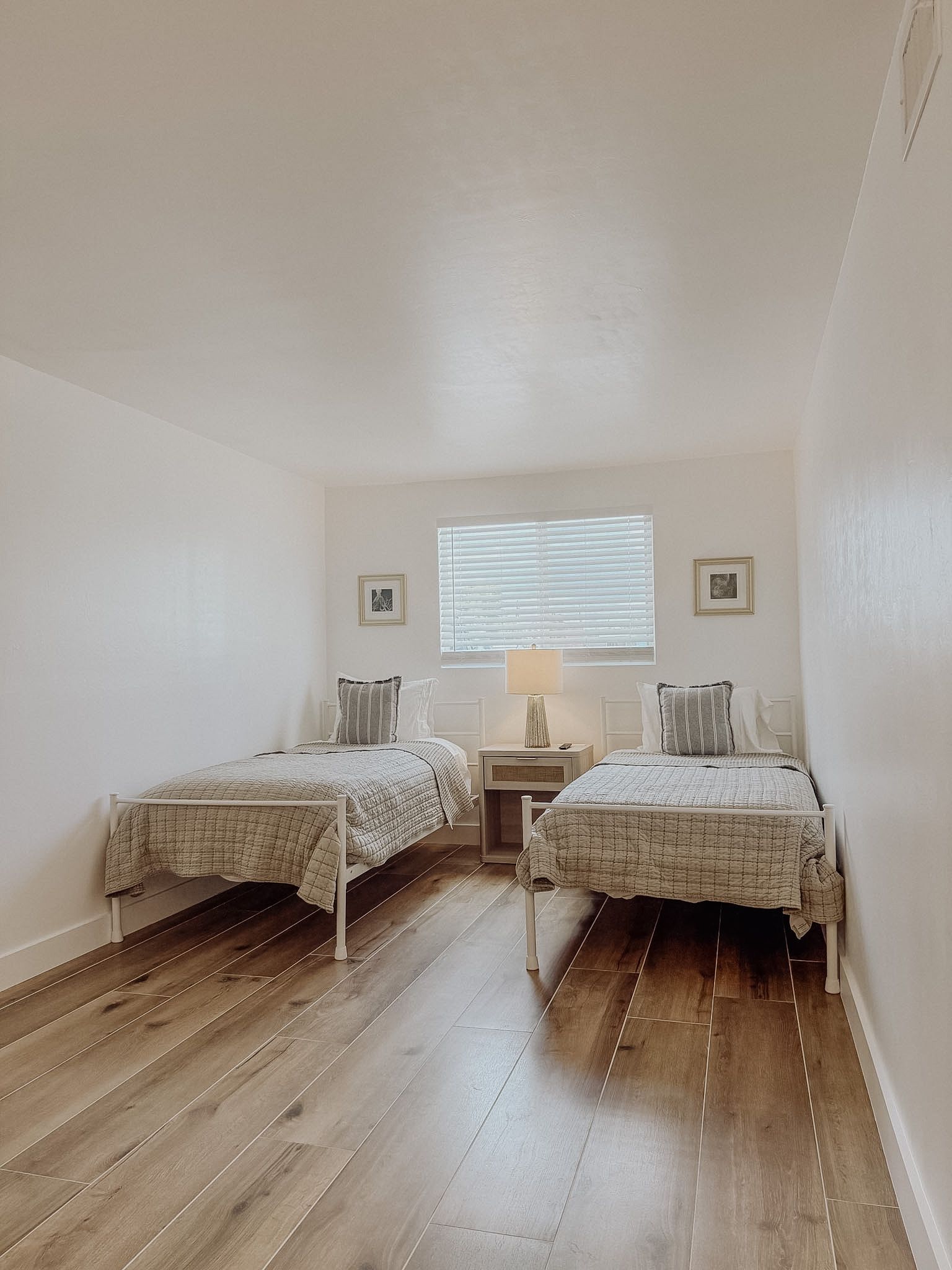 A bedroom with two single beds, a bedside table with a lamp, a window with blinds, and a framed picture on the wall.