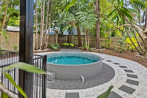 A circular hot tub surrounded by a pebble border, with a wooden fence and tropical plants in the background. - Tropical Koru Cottage - Private Pool - BBQ (Cairns)