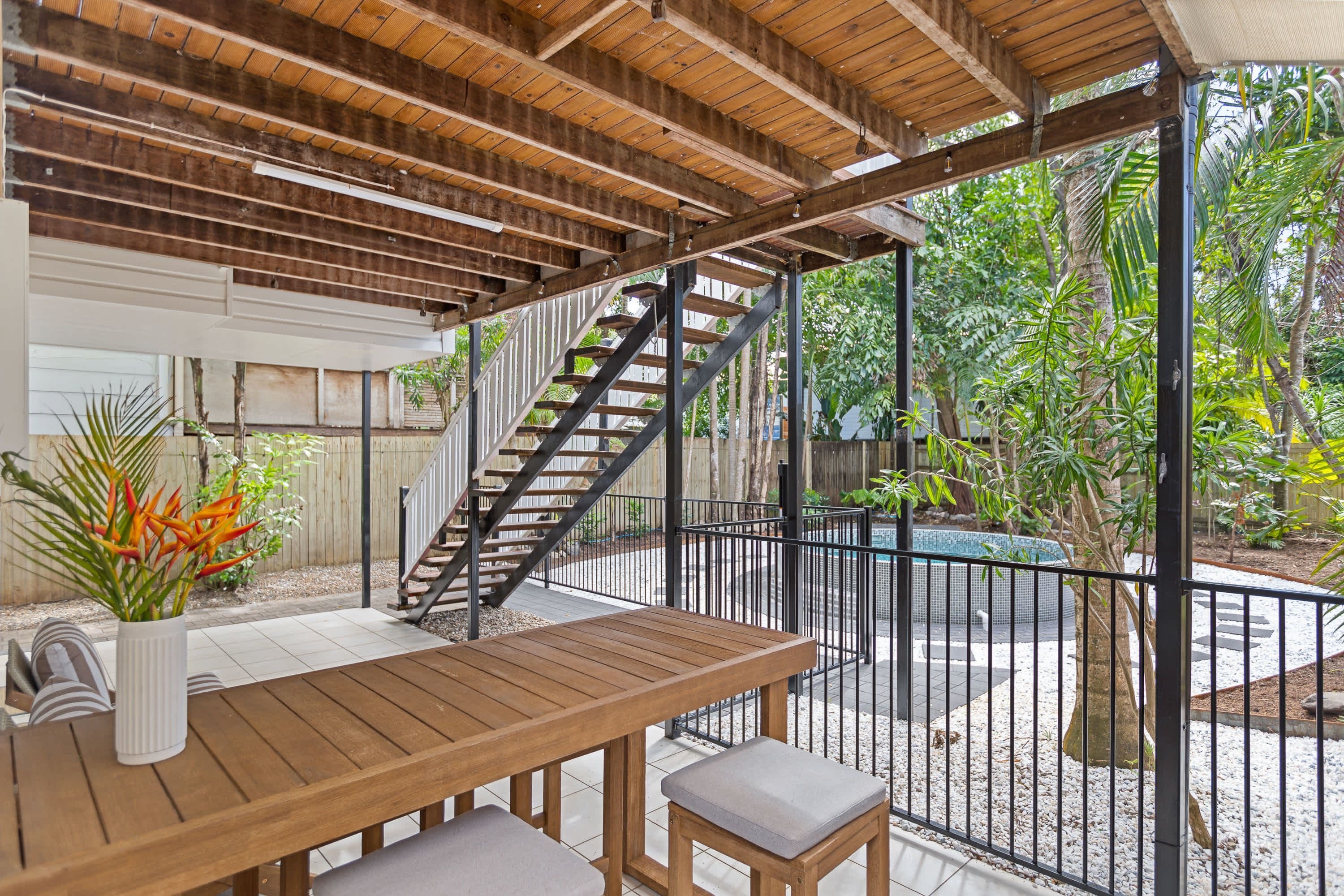 Área externa coberta com mesa e banco de madeira, escada e vista para um jardim com árvores e piscina.