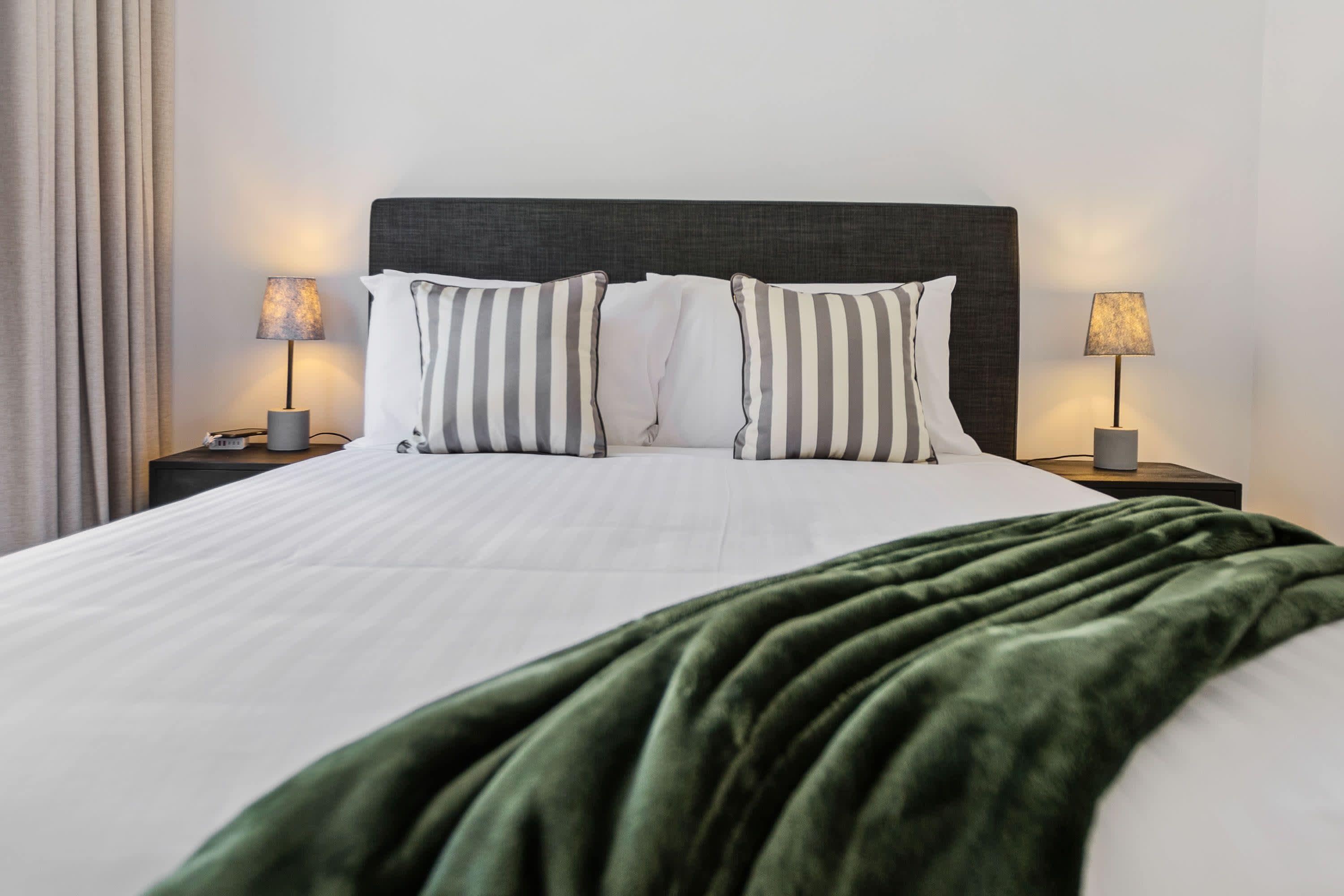 Una cama bien hecha con almohadas a rayas, cabecera oscura y manta verde.