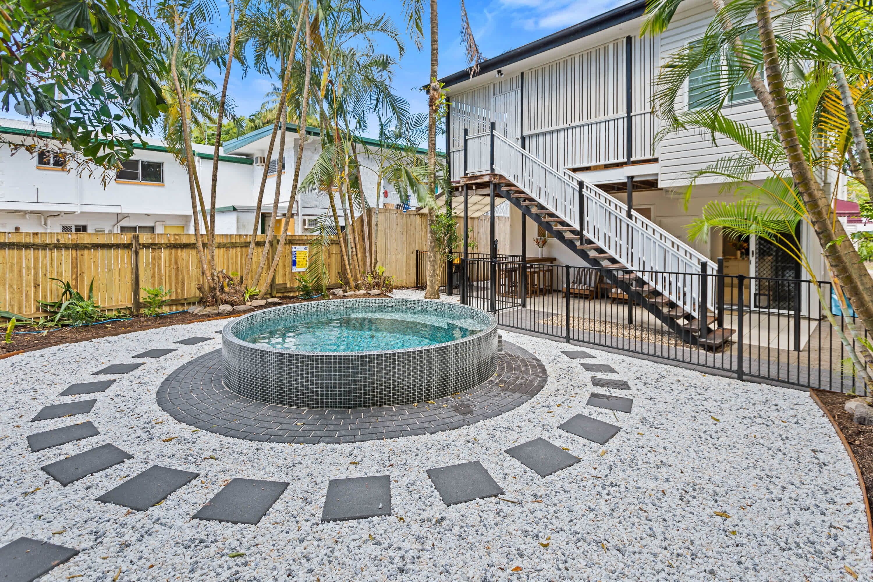 A circular hot tub surrounded by a pebble border, with a wooden fence and a staircase leading to a building.